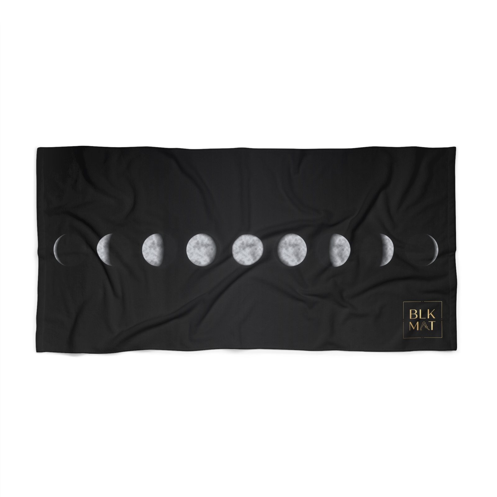 Black beach towel with a row of nine silver moon phases, new to full and back, across its middle and a small gold BLKMAT logo in the bottom-right corner.